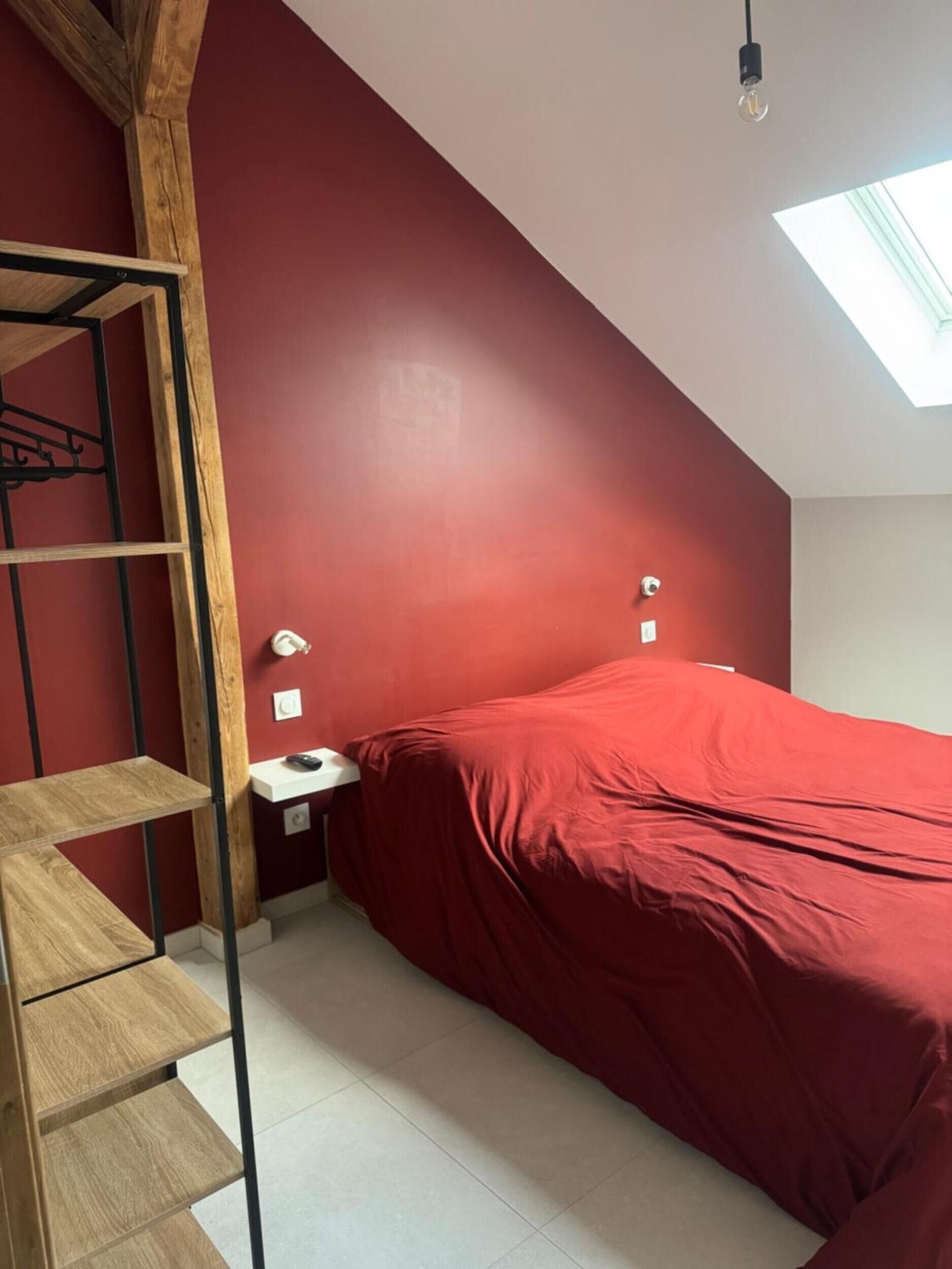 A bedroom with a red wall, a skylight, a bed with a red cover, a wooden ladder, and a nightstand with a lamp.