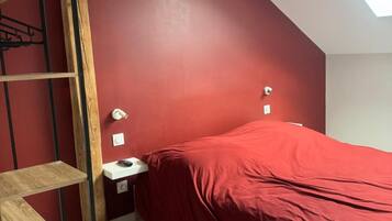 Un dormitorio con pared roja, tragaluz, cama con cubierta roja, escalera de madera y mesita de noche con lámpara.