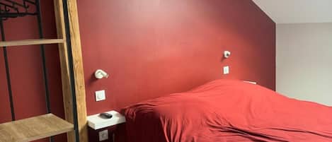 Ein Schlafzimmer mit roter Wand, Oberlicht, Bett mit roter Decke, Holzlader und Nachttisch mit Lampe.