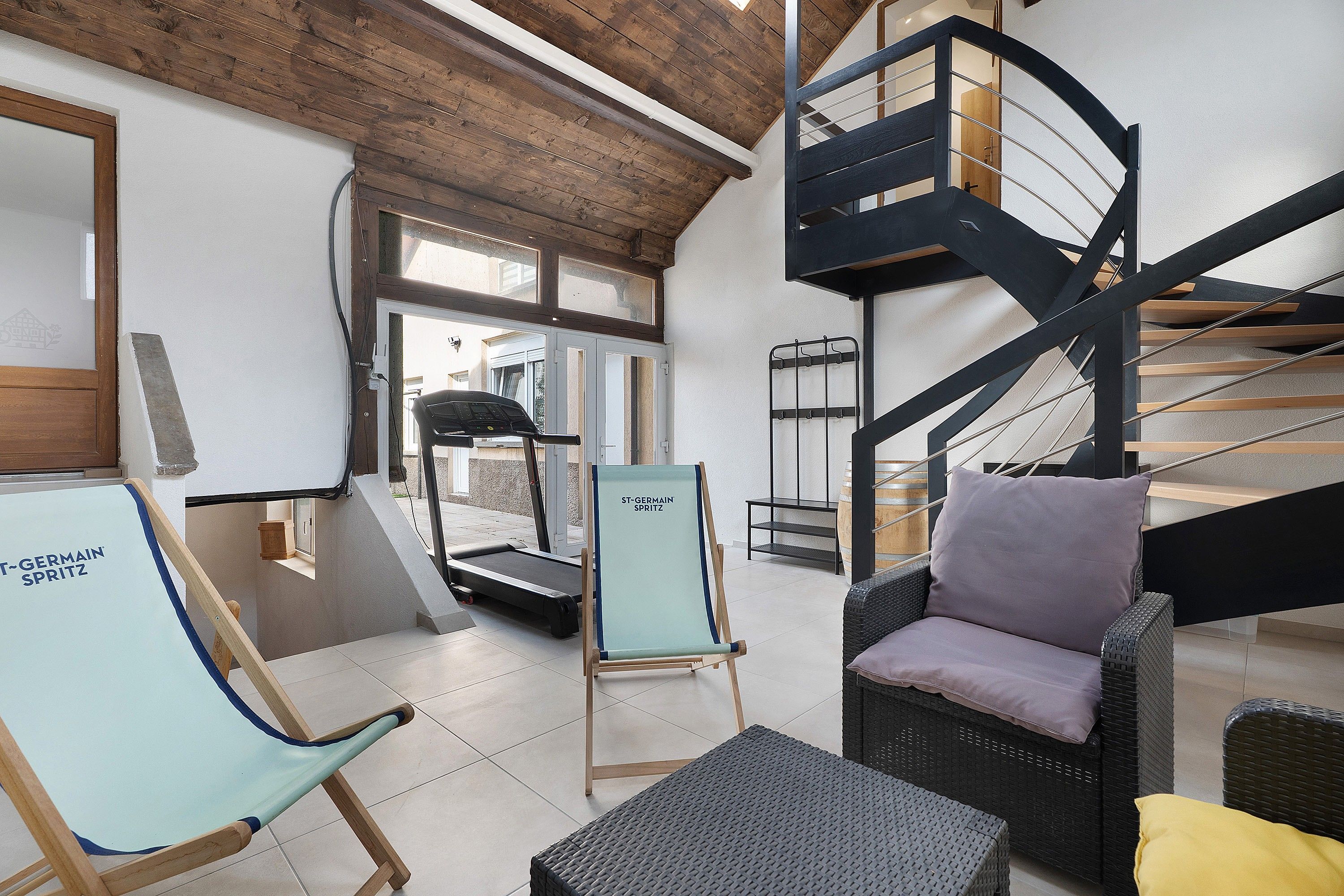 A modern interior with a treadmill, a blue exercise chair, a wicker sofa, and a black spiral staircase.