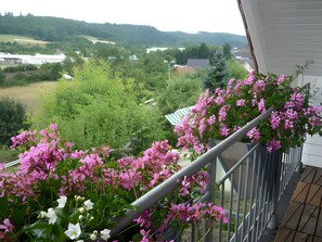 A balcony with pink flowers and a view of a green landscape with trees and buildings.