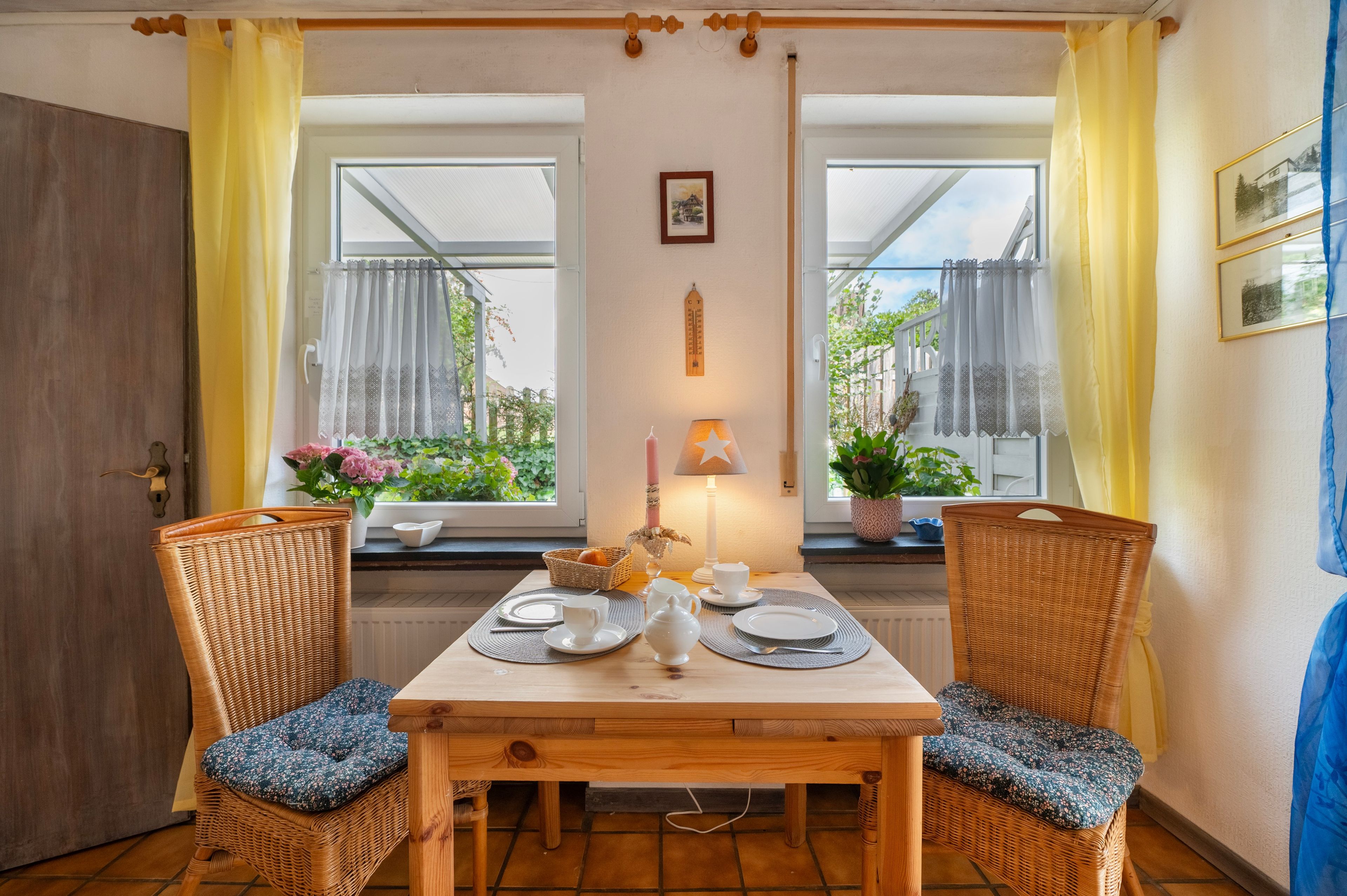 Un comedor con una mesa de madera y dos sillas de mimbre, una lámpara de mesa encendida, y vistas al verdor a través de las ventanas.