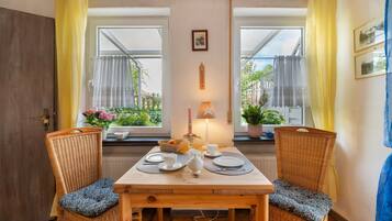 Un comedor con una mesa de madera y dos sillas de mimbre, una lámpara de mesa encendida, y vistas al verdor a través de las ventanas.