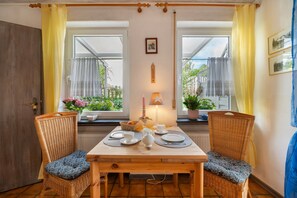 A dining area with a wooden table and two wicker chairs, a lit table lamp, and a view of greenery through the windows.