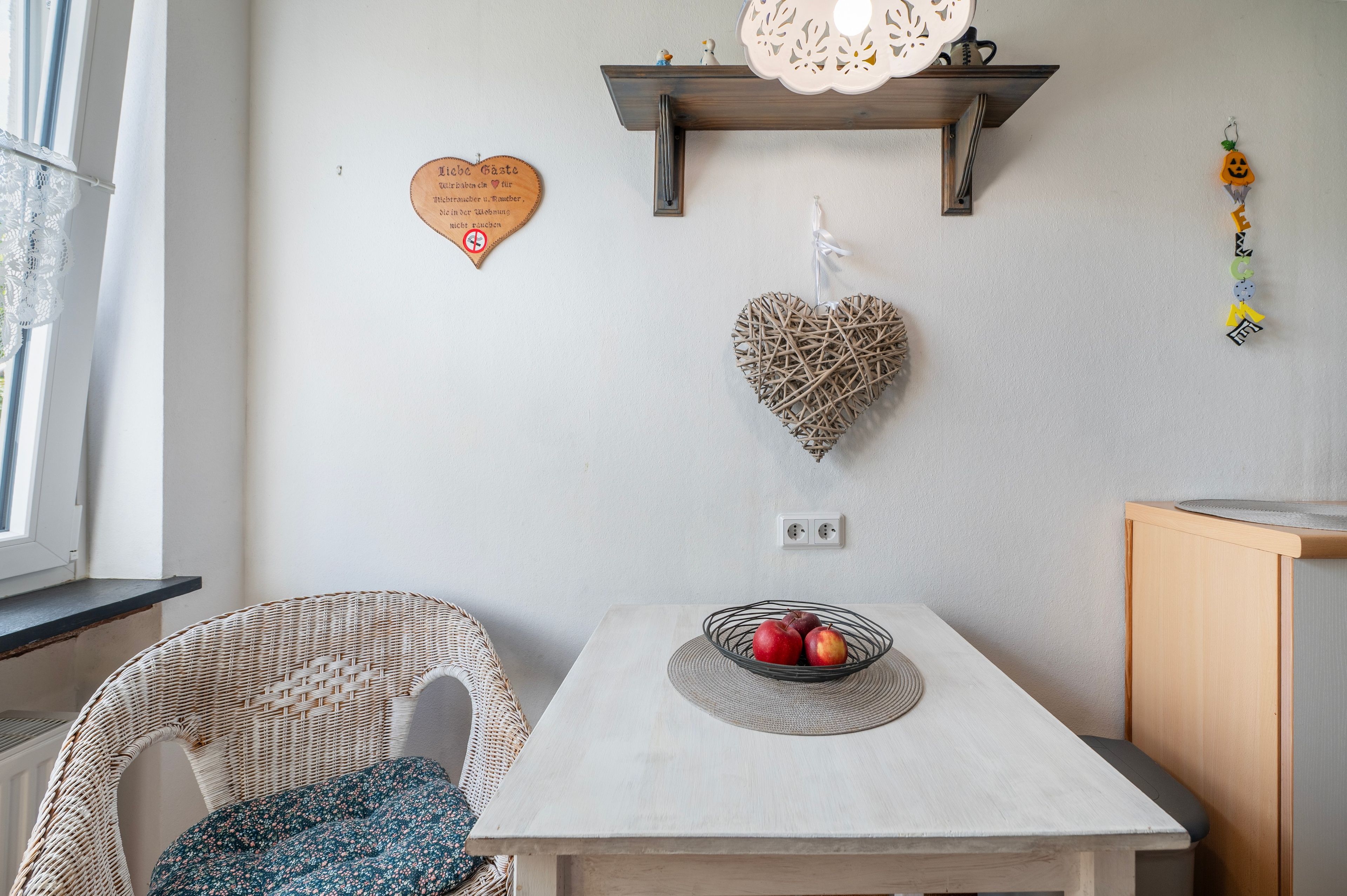 Ein Zimmer mit einem Tisch, Korbstühlen, einer Wanddekoration in Herzform und einem an der Wand befestigten Regal mit einem dekorativen Gegenstand.