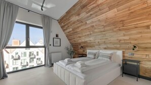 A bedroom with a sloped ceiling, a large bed, a bedside table, and a view of the city. - Granaria D601 | Balcony | AC | TOP Location (Gdańsk)