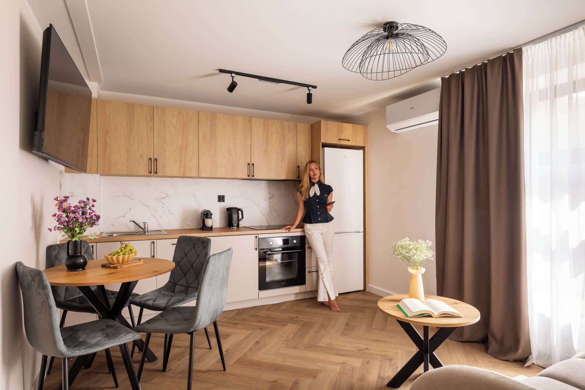 Eine moderne Küche mit Holdekor, ein Essbereich mit rundem Tisch und Stühlen, ein kleiner Beistelltisch mit einem Buch und ein großes Fenster mit Vorhängen.