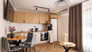 A modern kitchen with wooden cabinets, a dining area with a round table and chairs, a small side table with a book, and a large window with curtains.