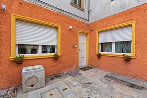 A courtyard with two windows, each with a yellow frame and a white shutter, and a door with a number plate.