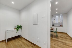 A room with a wooden floor, a white wall, a small table with a chair, a potted plant, and a window with a curtain.