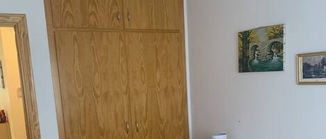 A bedroom with a wooden wardrobe, a bed with a patterned mattress, a bedside table with a lamp, and two wall-mounted paintings.