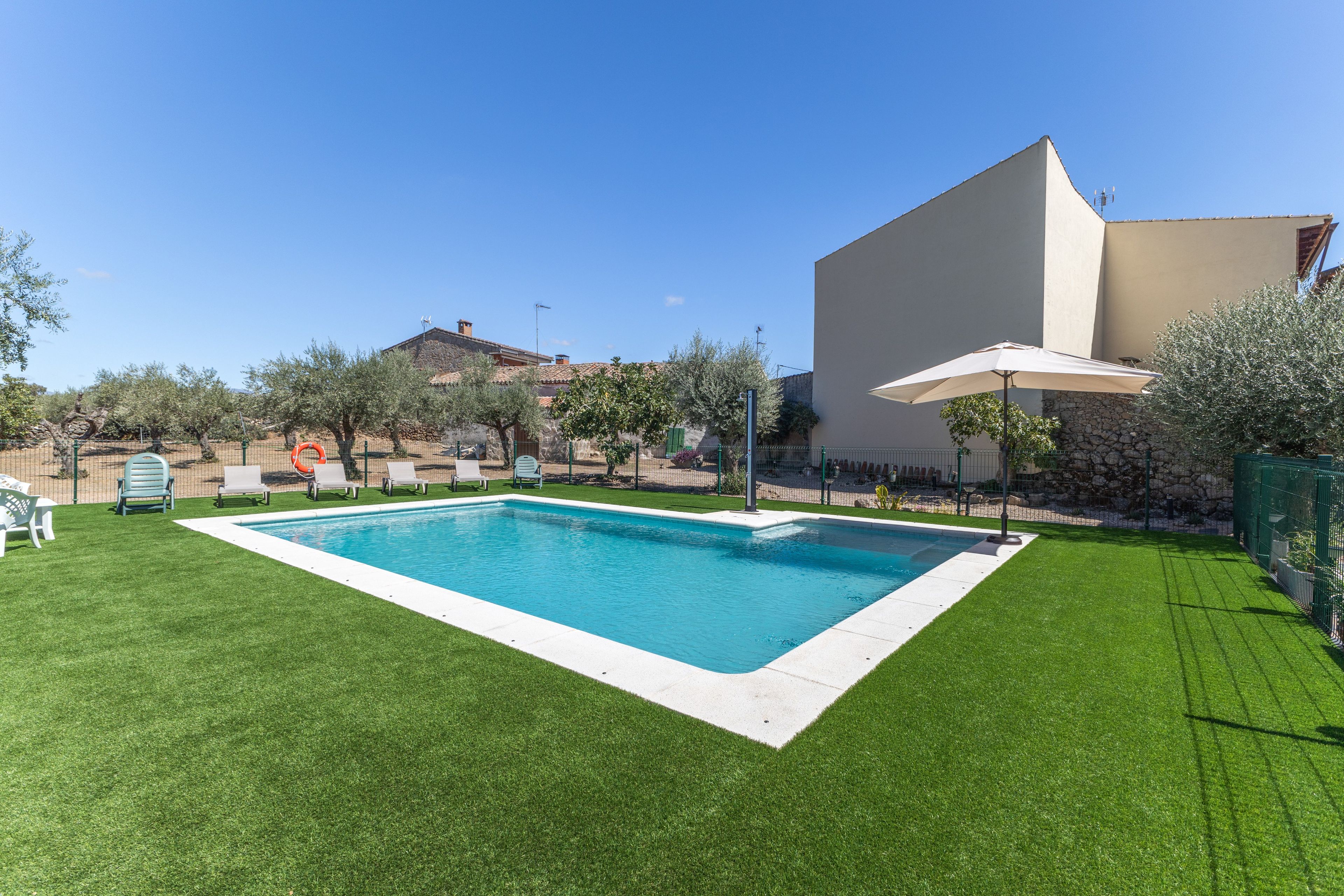 Une piscine rectangulaire bordée de blanc, entourée d’une pelouse bien entretenue, de quelques arbres et, au fond, d’un bâtiment dont le toit est en pente.