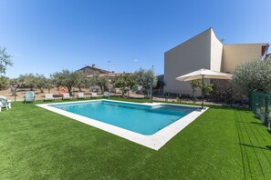 A rectangular swimming pool with a white edge, surrounded by a well-maintained lawn, a few trees, and a building with a sloped roof in the background. - Tourist Home 'La Posada del Pueblo', Sotillo de las Palomas (Sotillo de las Palomas)