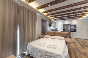 Quarto moderno com cama, vigas de madeira, cozinha ao fundo e porta do banheiro.