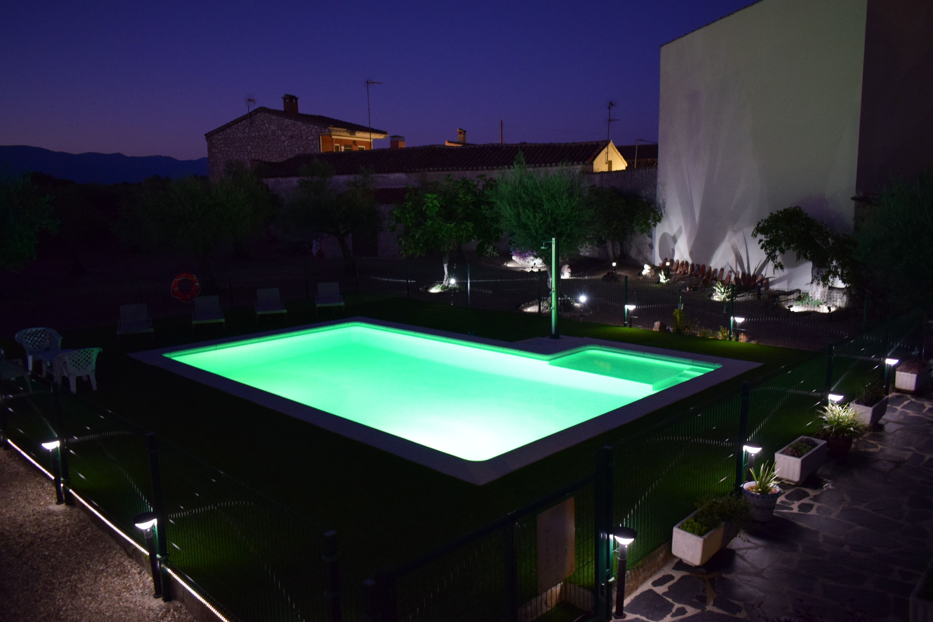 A rectangular swimming pool with a green interior, surrounded by a low wall and a fence. There are chairs and tables around the pool, and a building with a light on top is visible in the background.