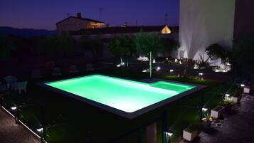 A rectangular swimming pool with a green interior, surrounded by a low wall and a fence. There are chairs and tables around the pool, and a building with a light on top is visible in the background.