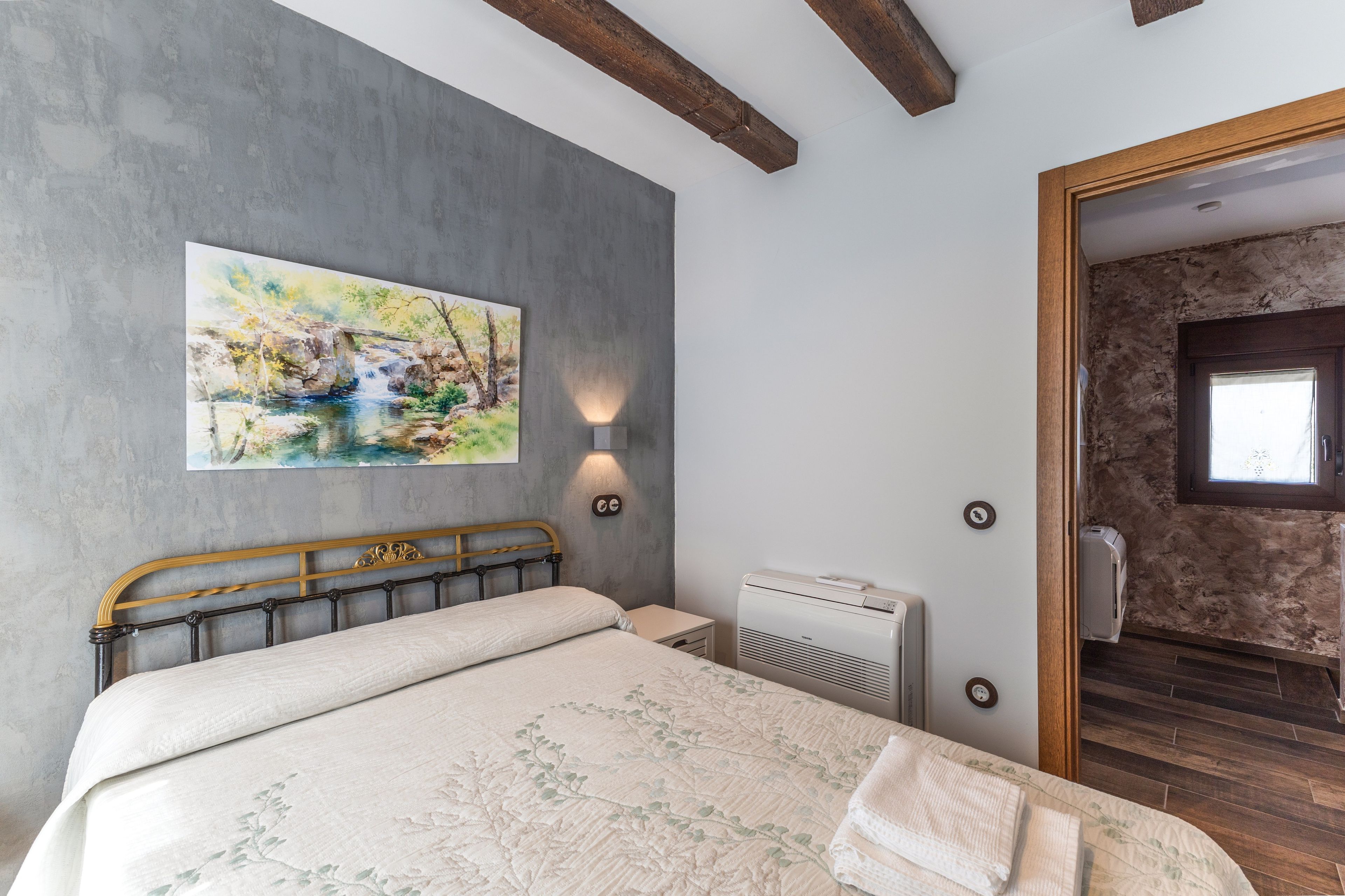 Een slaapkamer met een bed, een schilderij, een wandlamp, een airconditioningapparaat en een houten deur die naar een andere kamer leidt.