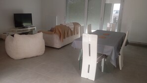 Un saló modern amb televisió de pantalla plana, un sofà, una taula de dinar amb caires i una finestra gran amb persianes.