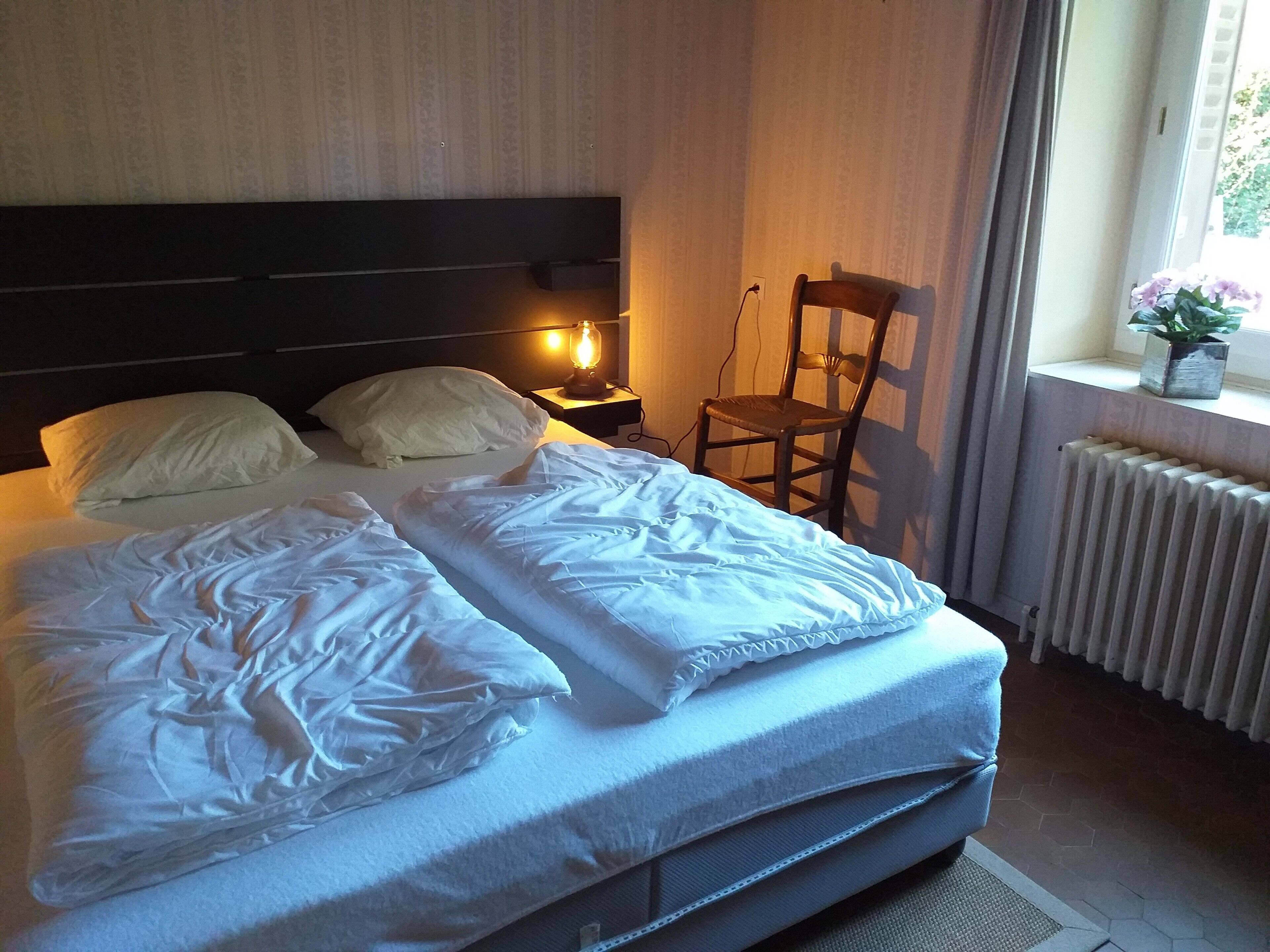 A hotel room with a bed, a chair, a nightstand with a lamp, and a radiator.