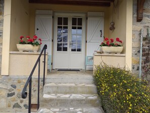 Una escalera de piedra conduce a una puerta blanca con contraventanas, macetas con flores rojas a cada lado y un pequeño porche.