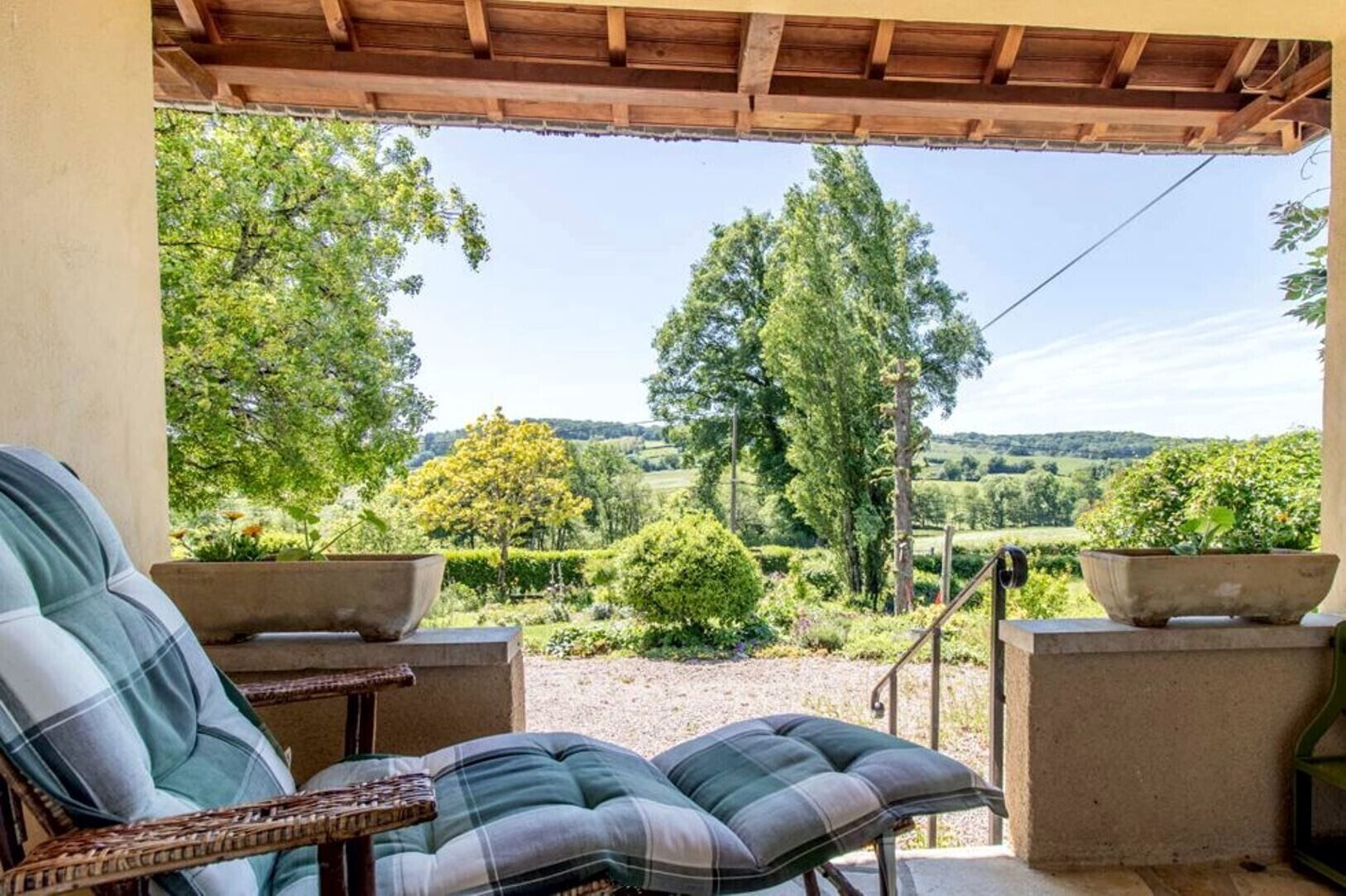 Een overdekt terras met twee stoelen en een tafel, met uitzicht op een weelderig landschap met bomen en een heldere lucht.