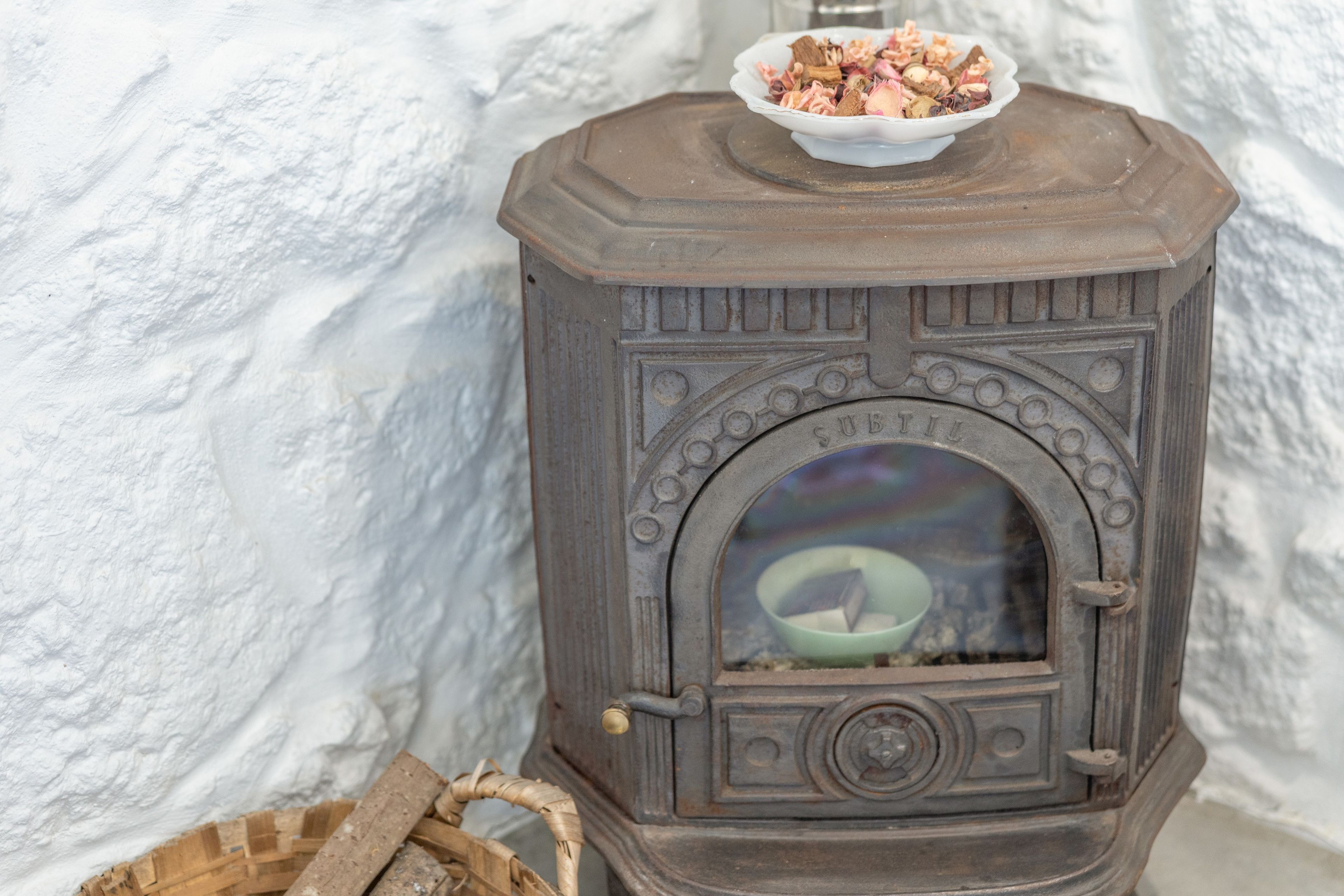 A wood-burning stove with a bowl of wood on top, a green bowl inside, and a basket of firewood beside it.