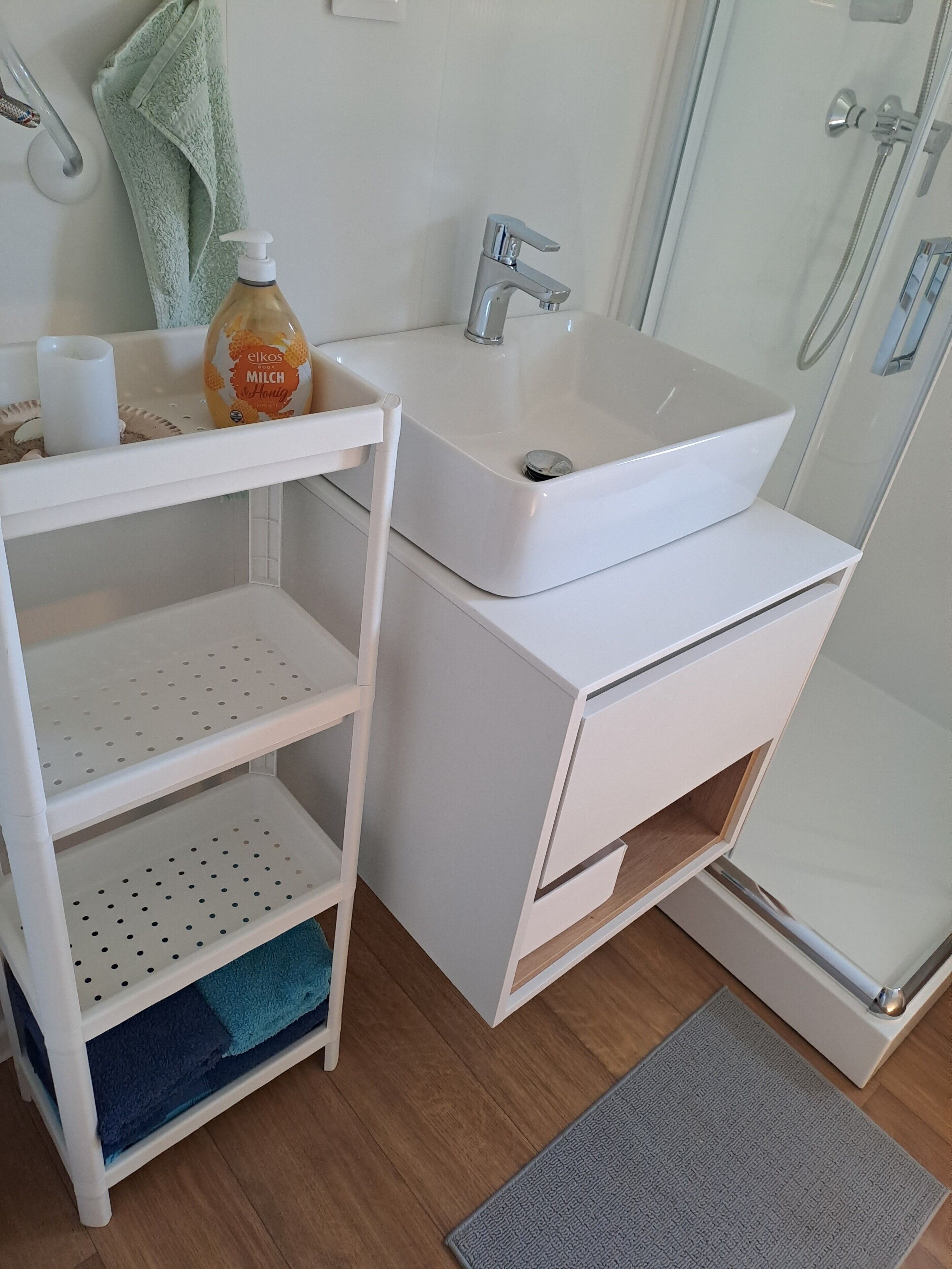 Un baño con lavamanos blanco, espejo, ducha y repisa con artículos de aseo.