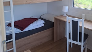 A bunk bed with a desk and chair, a window with a view of greenery, and a white lamp on the desk.