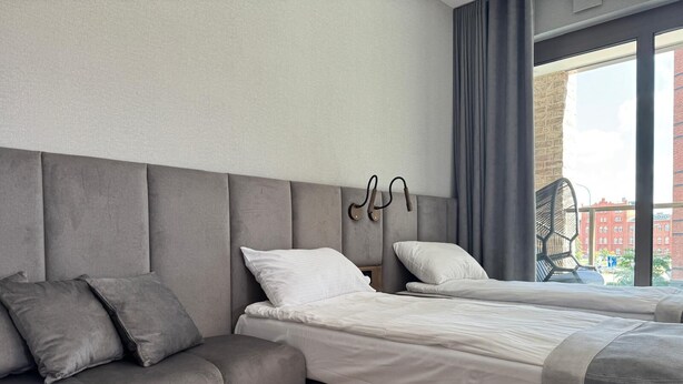 Ett modernt hotellrum med en grå soffa, två enkelsängar och utsikt över staden genom stora fönster.