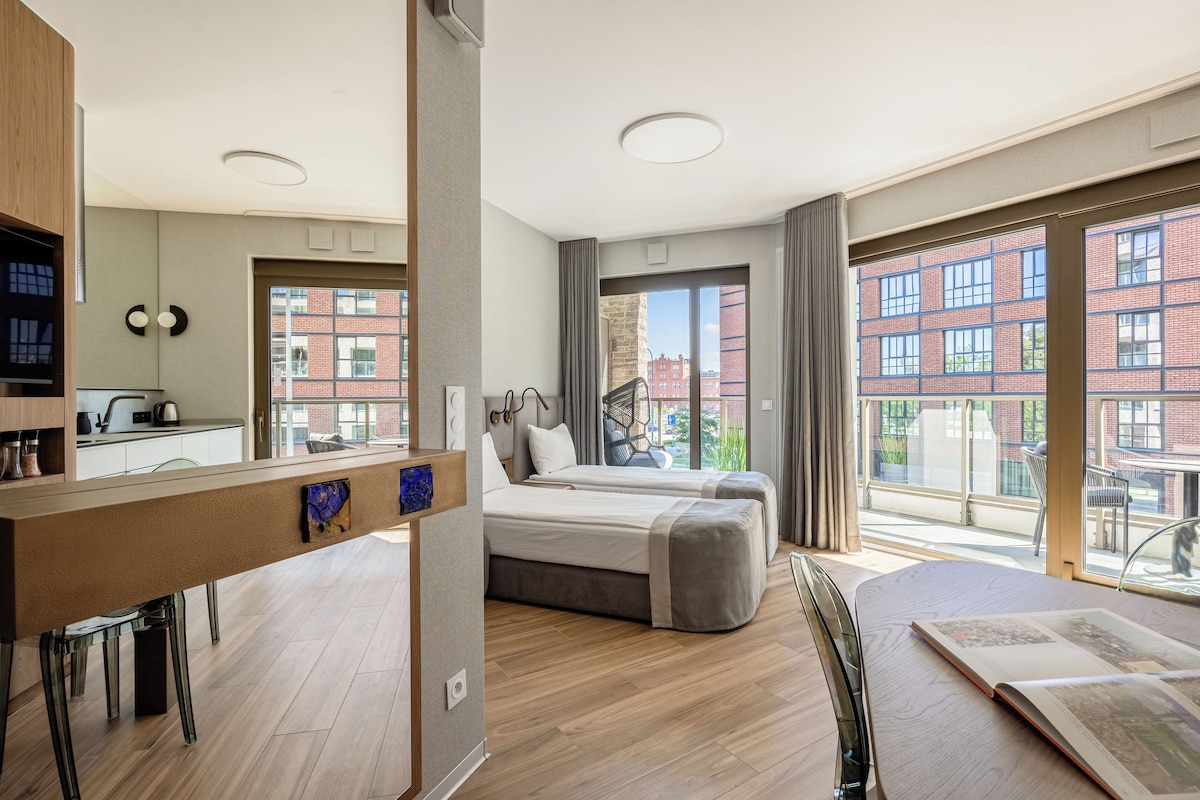 A modern hotel room with a kitchenette, a bed, a seating area, and large windows offering a view of the city.