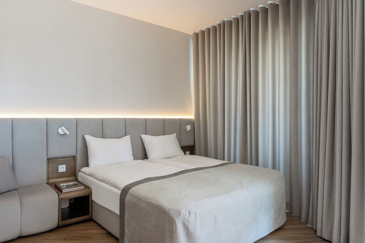 A modern hotel room with a large bed, a bedside table, a nightstand, and a wall-mounted lamp.