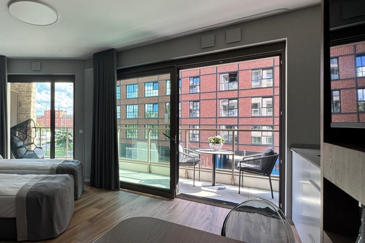 A hotel room with two beds, a balcony with a view of a red brick building, and a dining area with a table and chairs.