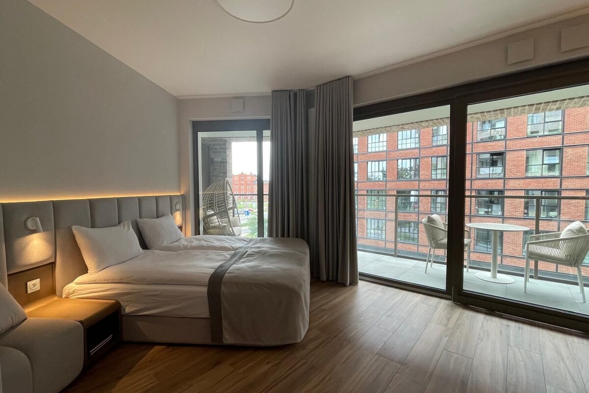 A modern hotel room with a large bed, a seating area, and a balcony with a view of a red brick building.