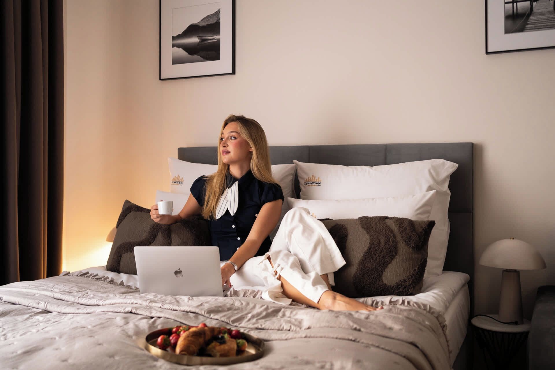 A person sitting on a bed with a laptop, holding a cup, and a plate of food.