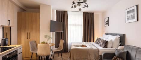 A hotel room with a bed, a sofa, a TV, a dining table with chairs, and a kitchen area.