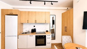 A compact kitchen with a refrigerator, oven, and coffee maker, a dining area with a table and chairs, and a TV mounted on the wall. - Kraków Bohaterów Września 8 | AC | Parkside (Kraków)