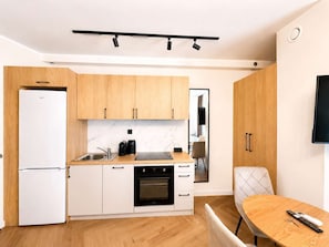 A compact kitchen with a refrigerator, oven, and coffee maker, a dining area with a table and chairs, and a TV mounted on the wall. - Kraków Bohaterów Września 8 | AC | Parkside (Kraków)