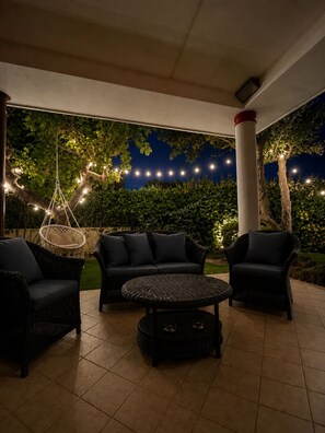 Een terras buiten met rieten meubilair, een hangstoel en lichtjes.