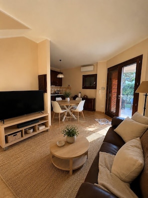 Una sala de estar con televisión de pantalla plana, un sofá, una mesa de centro con una planta y un comedor con sillas y una mesa.