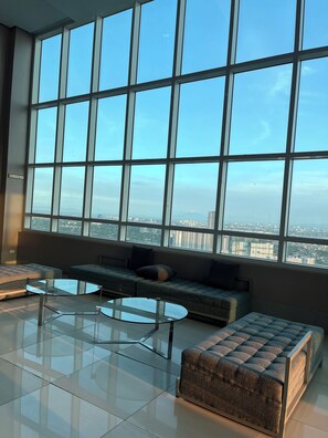 A modern lounge area with large windows, a sofa, a glass coffee table, and a cushioned ottoman. - Maison La Giada  Cozy 2BR Staycation in Makati (Makati)