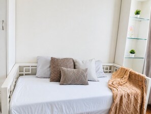 Ein Bett mit weißer Bettwäsche und mehreren Kissen, ein Glastischchen mit Pflanzen und eine weiße Wand.