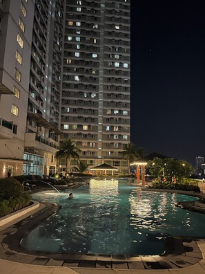 A nighttime view of a hotel with a swimming pool, a tall building in the background, and a covered area with palm trees. - Maison La Giada  Cozy 2BR Staycation in Makati (Makati)