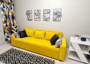 Un sofá amarillo brillante con varios almohadones, una alfombra estampada y una mesita auxiliar blanca con una planta.