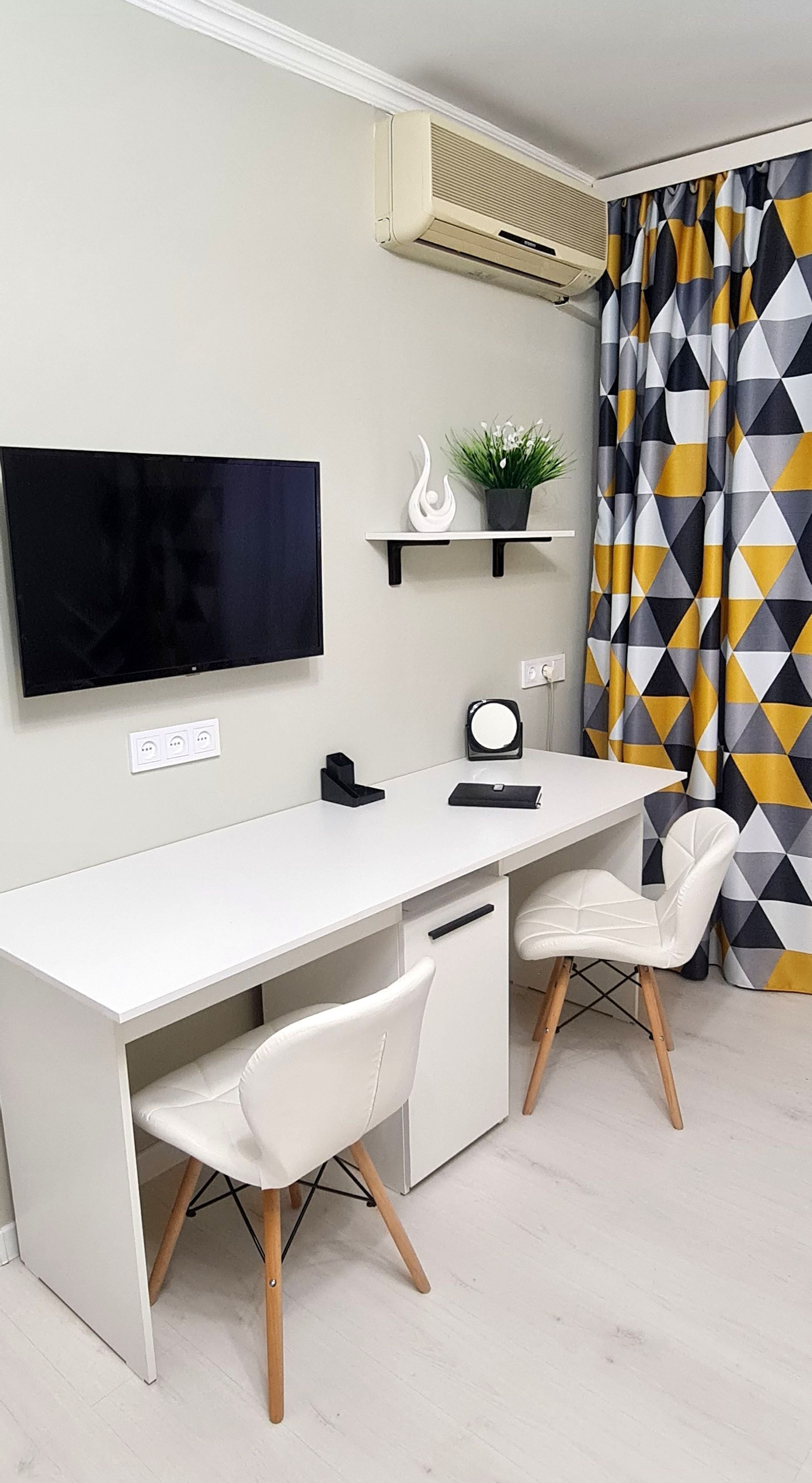 Habitació moderna i minimalista amb escriptori blanc, dues cadires blanques, televisió penjada, petita prestatgeria amb objectes decoratius i cortina amb dibuixos geomètrics.