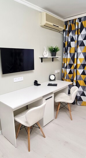 Habitación moderna y minimalista con un escritorio blanco, dos sillas blancas, un televisor montado, un estante con objetos decorativos y una cortina con estampado geométrico.