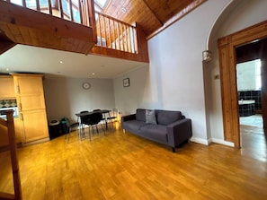 A spacious living area with a wooden ceiling, a gray sofa, a dining table with chairs, and a kitchen area with cabinets.