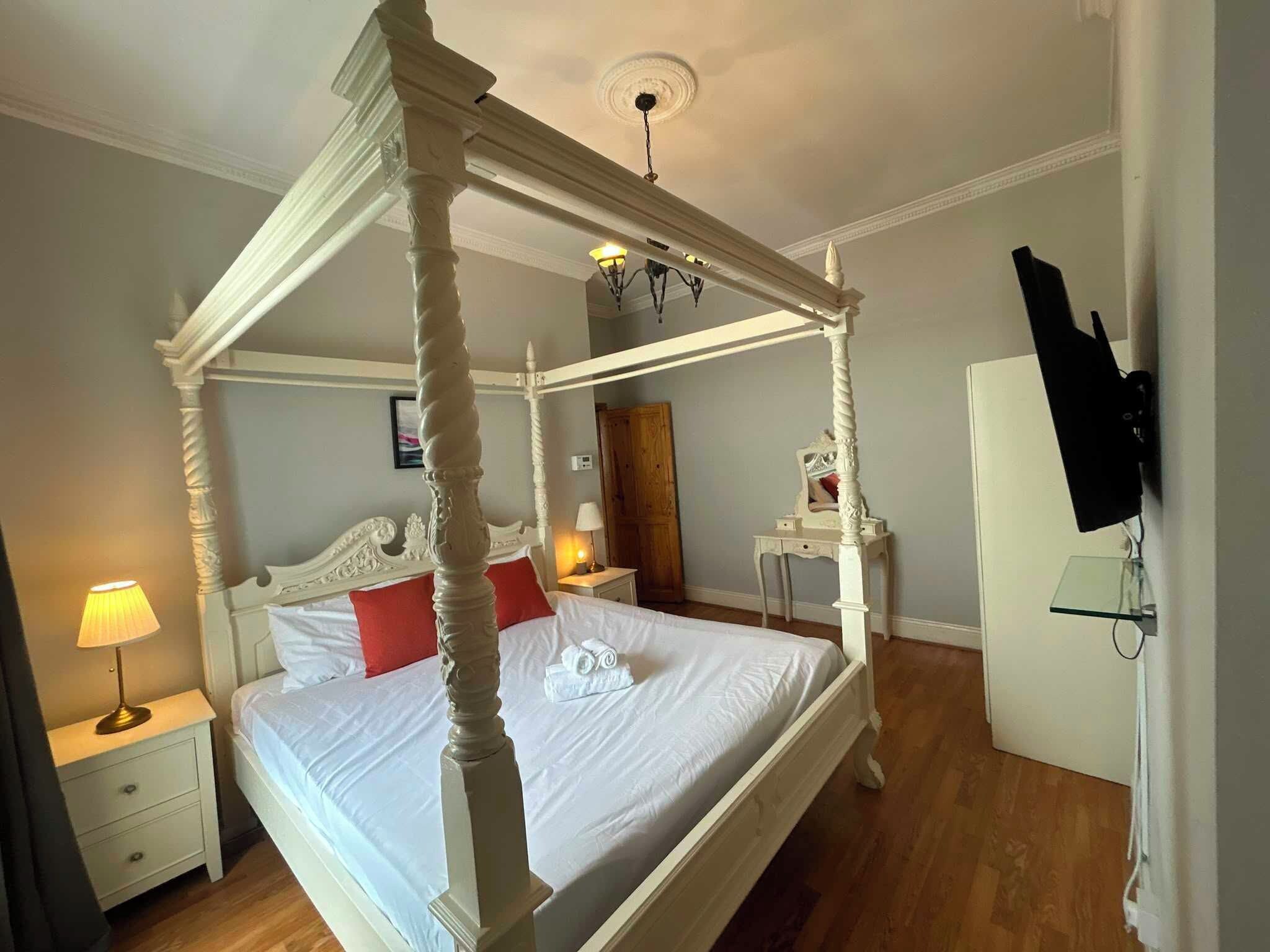 A four-poster bed with white bedding, a bedside table with a lamp, and a wooden floor.