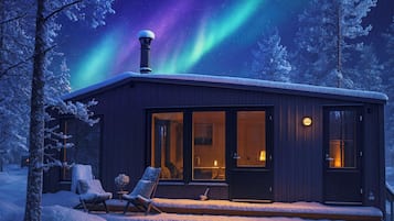 En hytte med en glasdør, en skorsten og en snedækket terrasse med to stole.