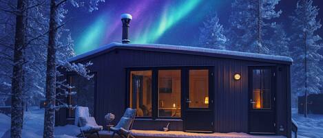 Une cabane avec une porte en verre, une cheminée et une terrasse enneigée équipée de deux chaises.