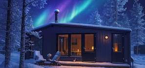 A cabin with a glass door, a chimney, and a snow-covered deck with two chairs.